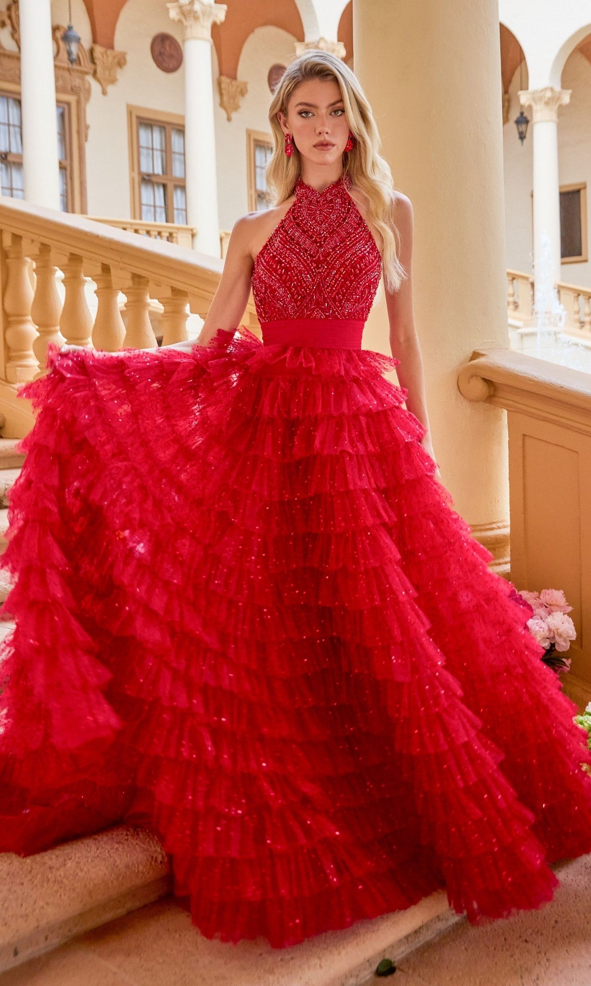 A woman wearing the Ava Presley 47258 long red prom dress with a sleeveless beaded halter bodice and tiered full skirt stands gracefully on a stone staircase before a classical building.